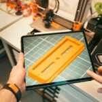 A person uses a tablet to view a 3D model prototype in a workshop equipped with 3D printers.